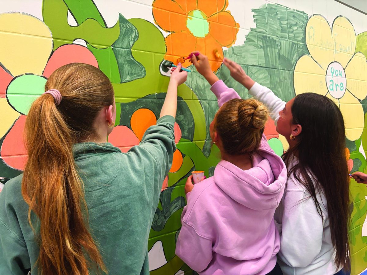 One flower at a time…On April 21, (from left) freshmen Eleanor Brewer, Chloe Culen and Ella Shipon paint the Words on Worth mural located in the high school’s main concourse. 